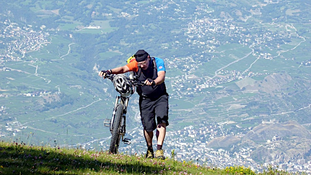 Ein Biker schiebt sein Rad sehr steil hoch. Weit unten im Tal sieht man Ortsschaften