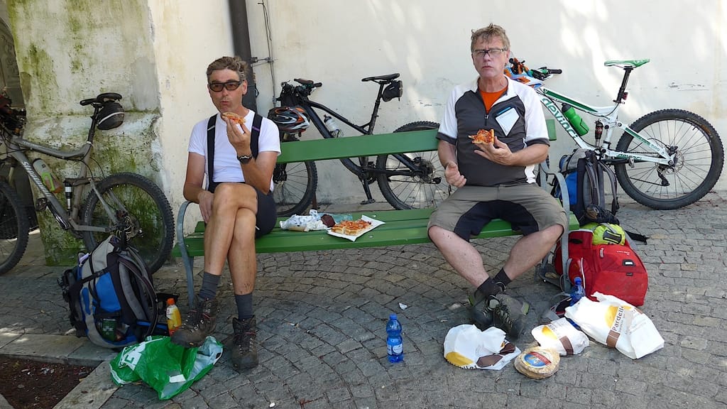 Eine Bank am Marktplatz in Sterzing. Zwei Biker essen mit offensichlichem Hunger Pizza